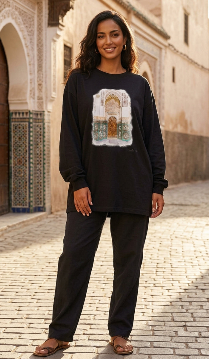 A woman stands outdoors in a sunlit street wearing a black long-sleeve top with a large rectangular architectural graphic on the front and black pants.