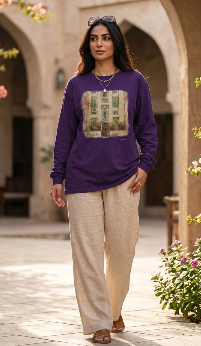 A woman wearing a purple long-sleeve crewneck sweatshirt with a large graphic print of a green garden door, paired with beige linen trousers, walking in a sunlit courtyard with arches.