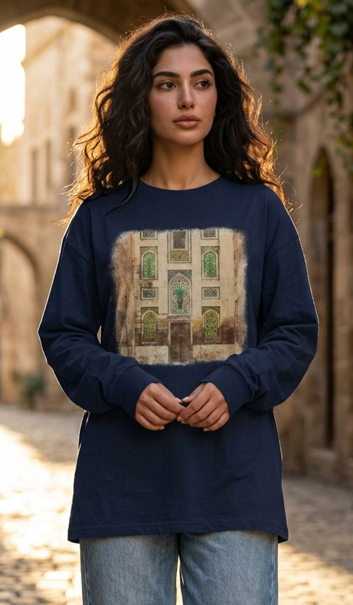 A woman wearing a navy blue long-sleeve t-shirt with a large rectangular graphic print of a traditional Middle Eastern building facade, set against a blurred stone archway background.