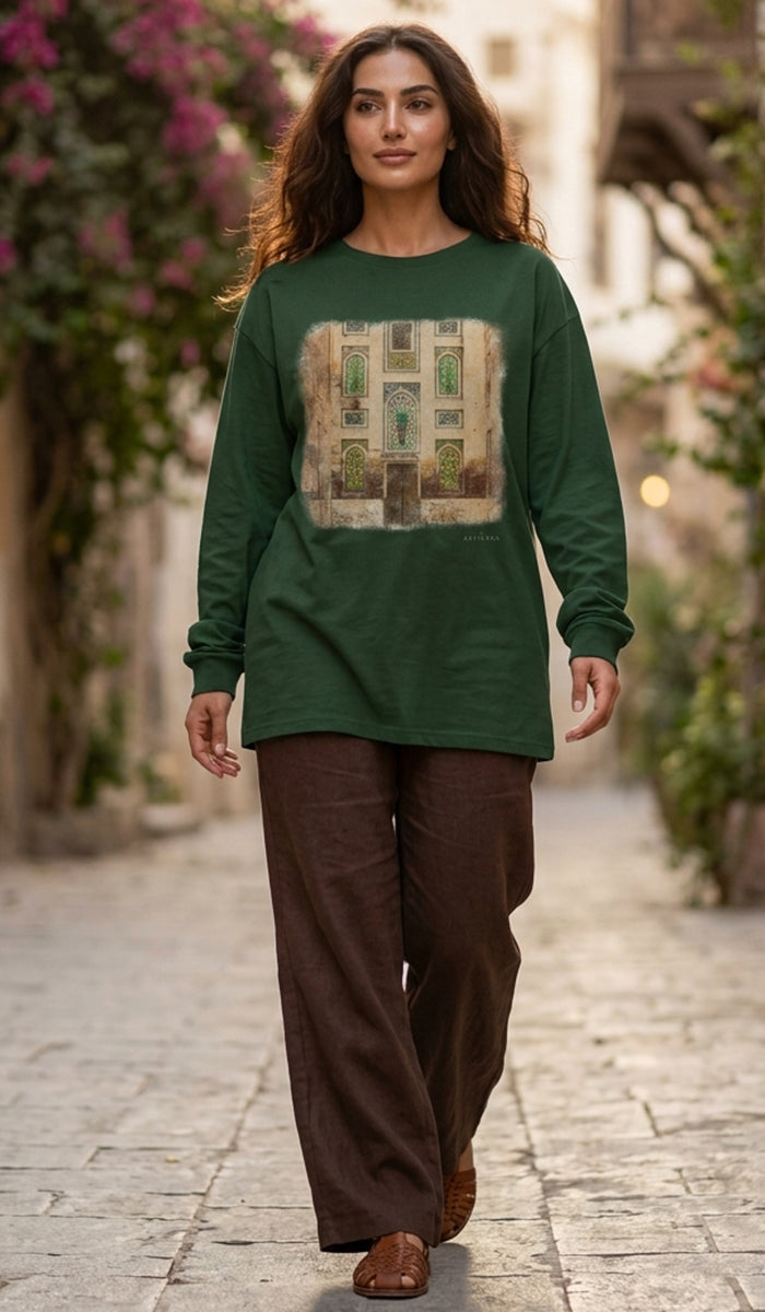 A woman walking down a cobblestone street wearing a dark green long-sleeve tunic top with a large vintage-style building graphic on the front, paired with brown wide-leg pants.