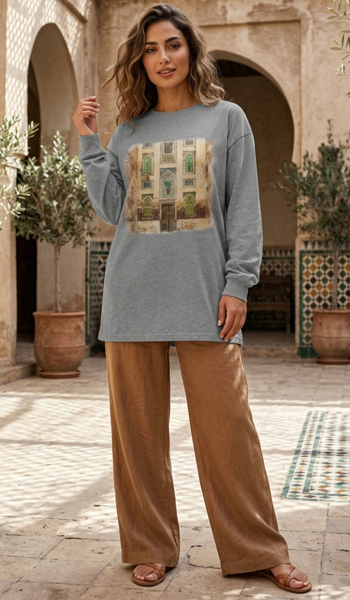 A woman stands in a sunlit courtyard wearing a loose heather gray long-sleeve top with a large multicolor architectural graphic on the front, paired with wide-leg tan pants and brown sandals.