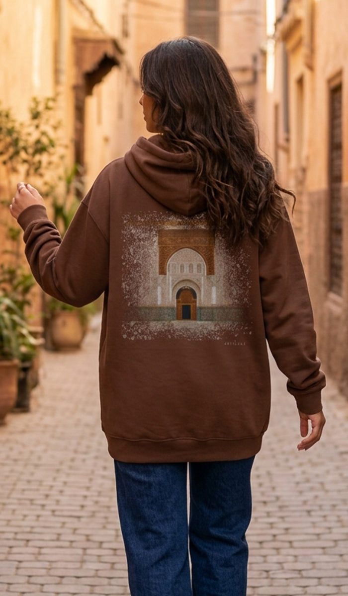 A woman viewed from behind wearing a brown pullover hoodie with a large printed graphic of a historic archway on the back, paired with blue jeans, walking down a sunlit cobblestone alley.