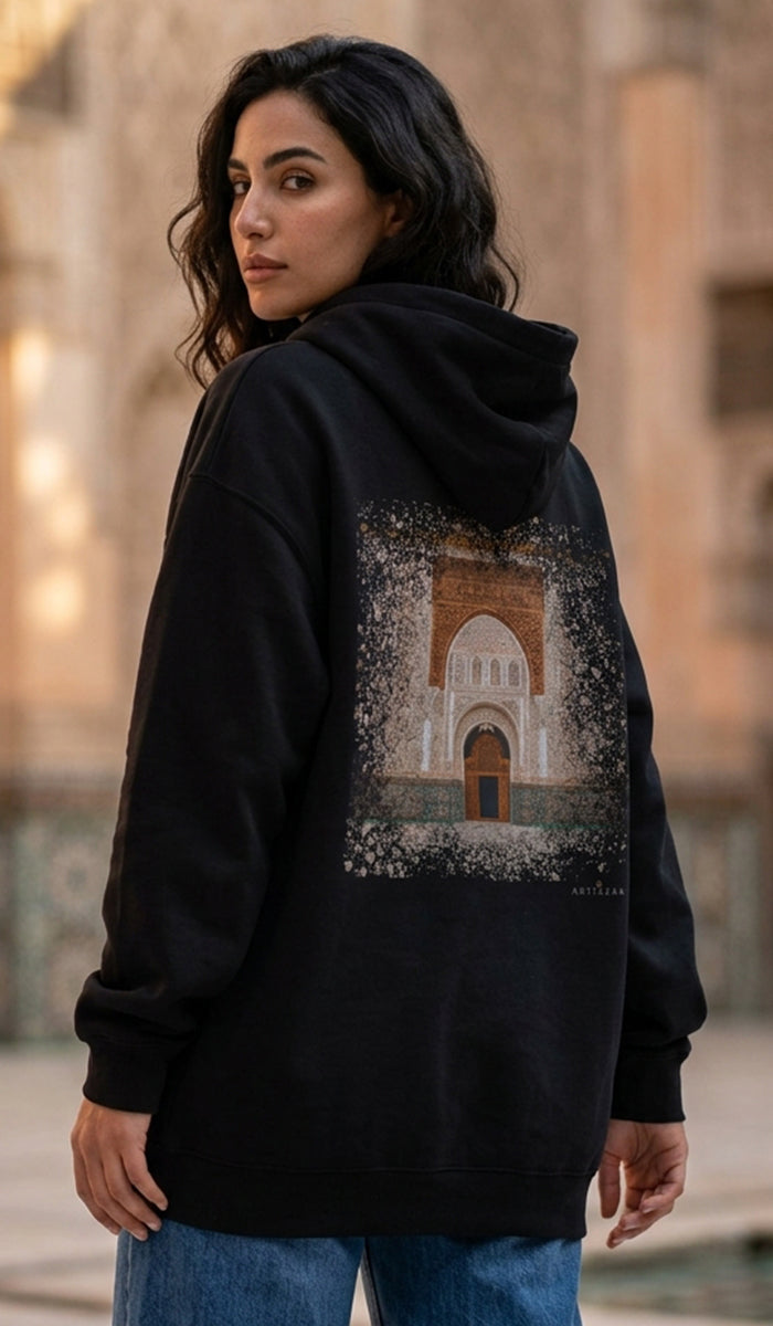 A woman wearing a black pullover hoodie with a large graphic print of an arched stone building on the back, photographed outdoors against a blurred stone building background.