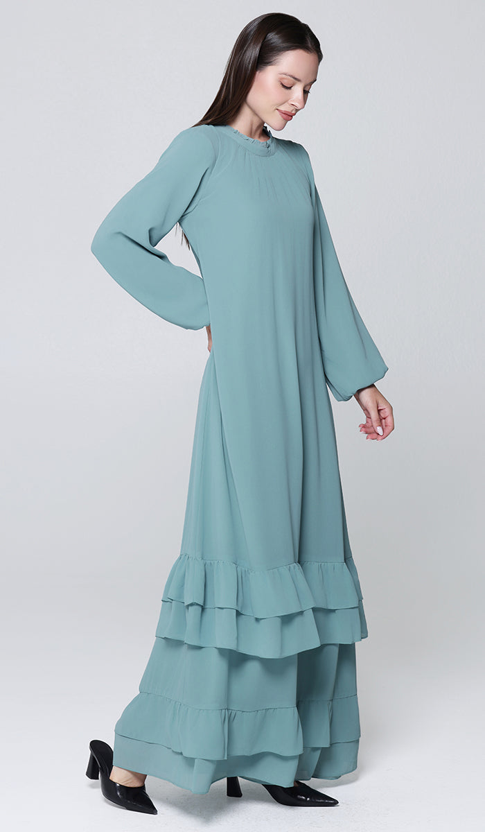 Woman wearing a long teal dress with ruffled layers on a white background