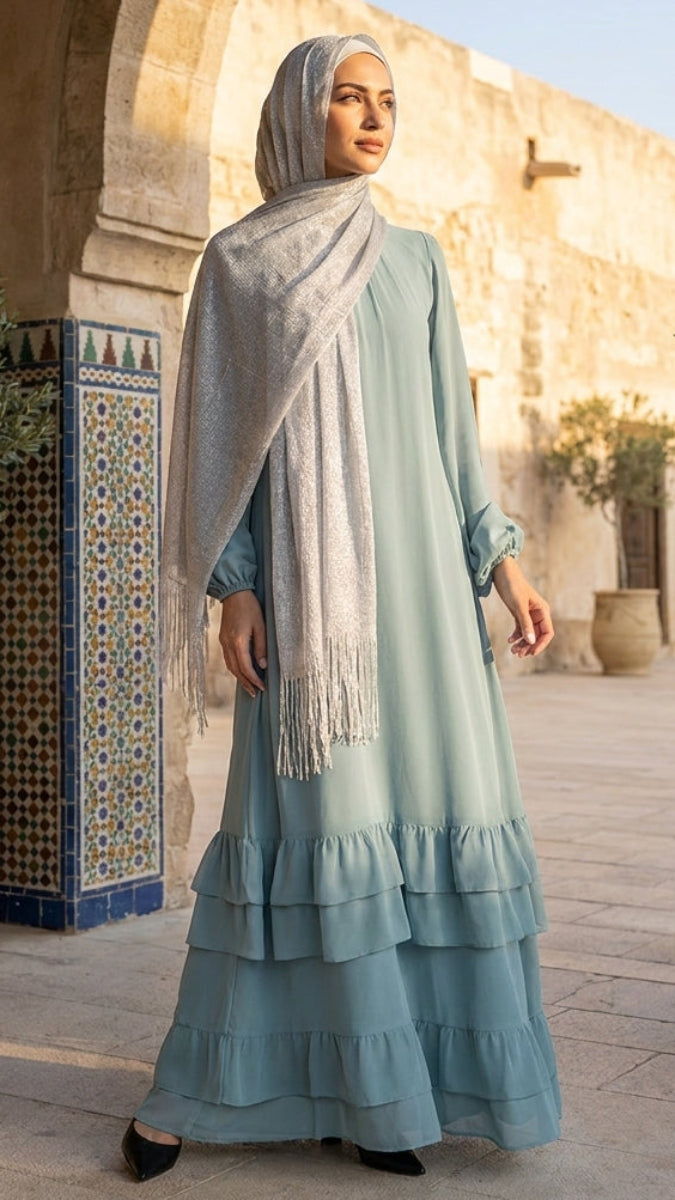 Woman in a light blue dress and headscarf standing in a sunlit courtyard with architectural elements.
