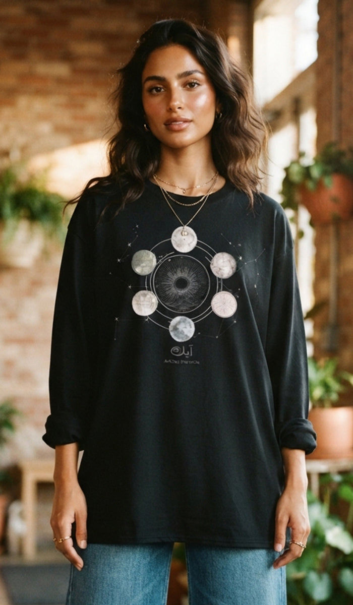 A woman wearing a black long-sleeve graphic t-shirt with a moon phase design on the front, paired with blue jeans, standing in a rustic indoor setting with brick walls and plants.