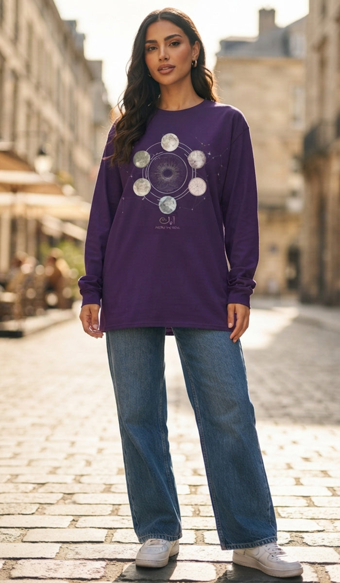 A woman stands on a cobblestone street wearing a purple long-sleeve graphic sweatshirt with a moon phase design, blue jeans, and white sneakers.