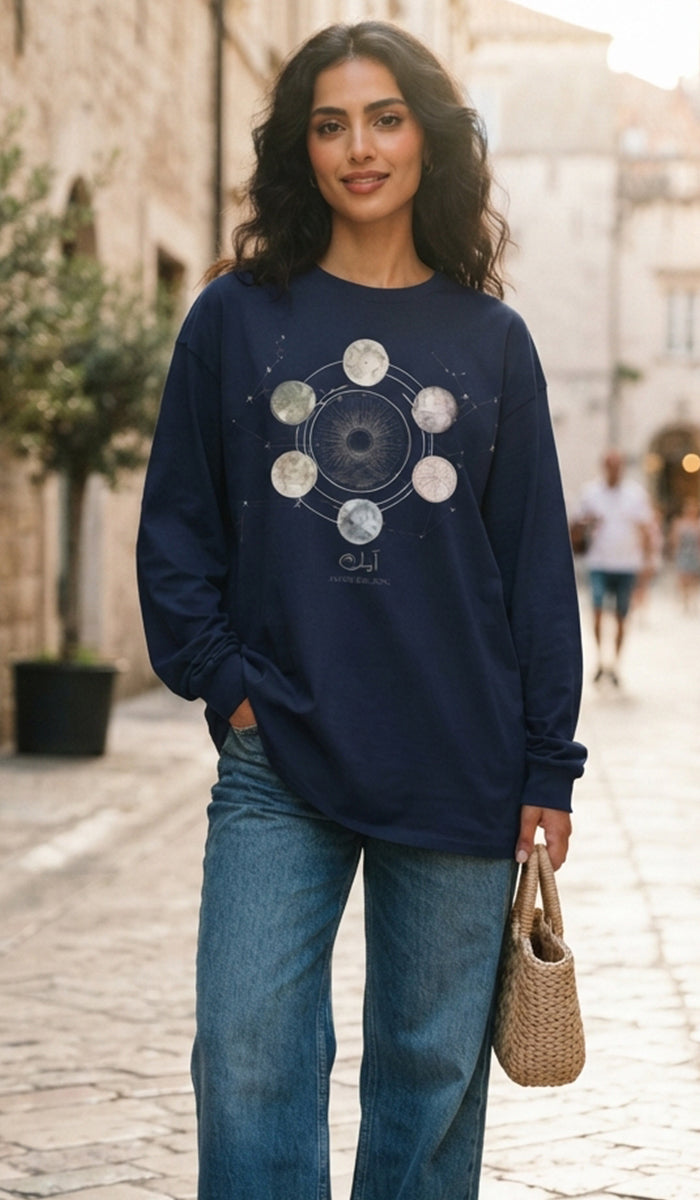 A woman wearing a navy blue long-sleeve crewneck sweatshirt with a moon phase graphic on the front, paired with blue jeans and holding a woven straw bag, standing in an outdoor street setting.