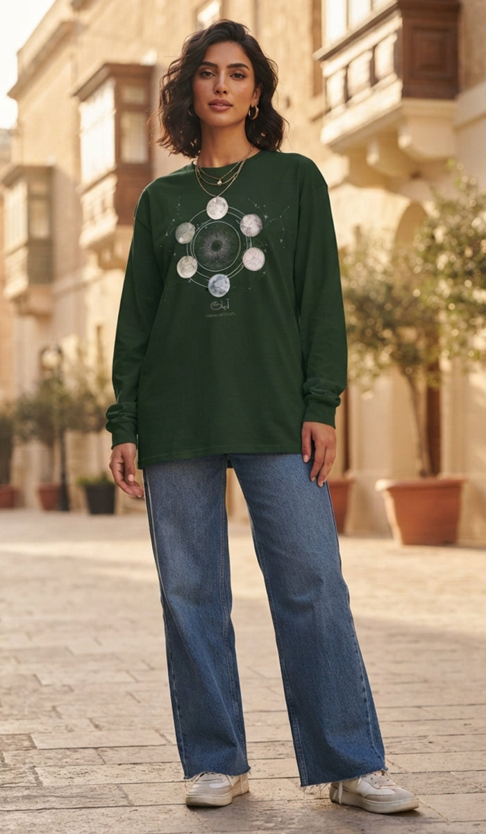 A woman stands on a sunlit cobblestone street wearing a dark green long-sleeve graphic sweatshirt with a circular sunburst design on the front, blue wide-leg jeans, and white sneakers. The background shows warm-toned buildings and potted plants.
