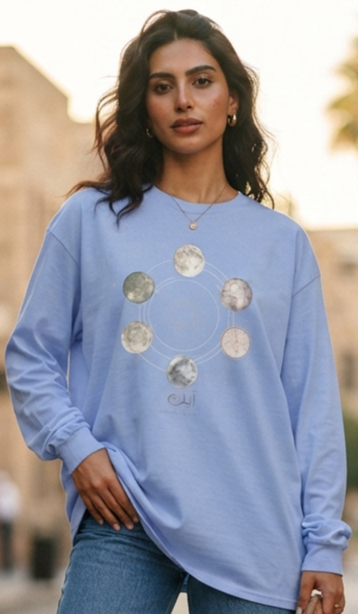 A woman wearing a light blue long-sleeve t-shirt with a circular graphic of metallic silver coins, paired with blue jeans, set against a blurred outdoor background.
