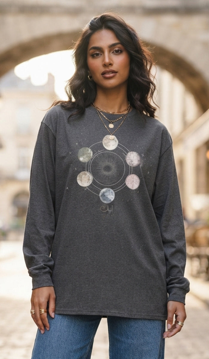 A woman wearing a dark gray long-sleeve t-shirt with a celestial moon phase graphic on the front, paired with blue jeans, standing in front of a blurred stone archway background.
