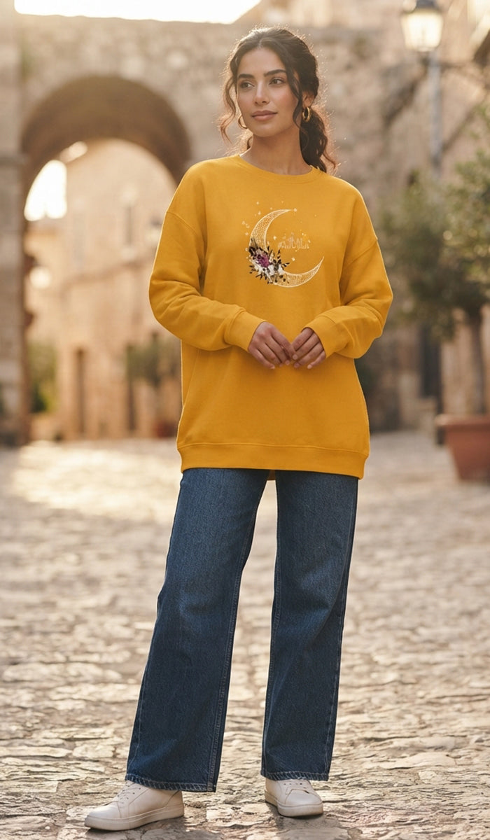 A woman stands on a cobblestone street wearing a mustard yellow sweatshirt with a moon and stars graphic, blue jeans, and white sneakers.