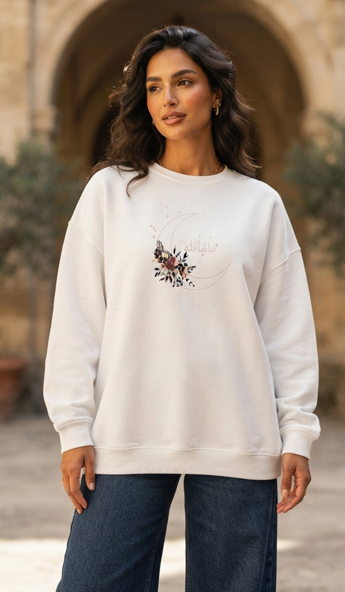 A woman wearing a cream-colored crewneck sweatshirt with a floral graphic on the chest, paired with dark blue jeans, standing in front of a blurred stone archway background.