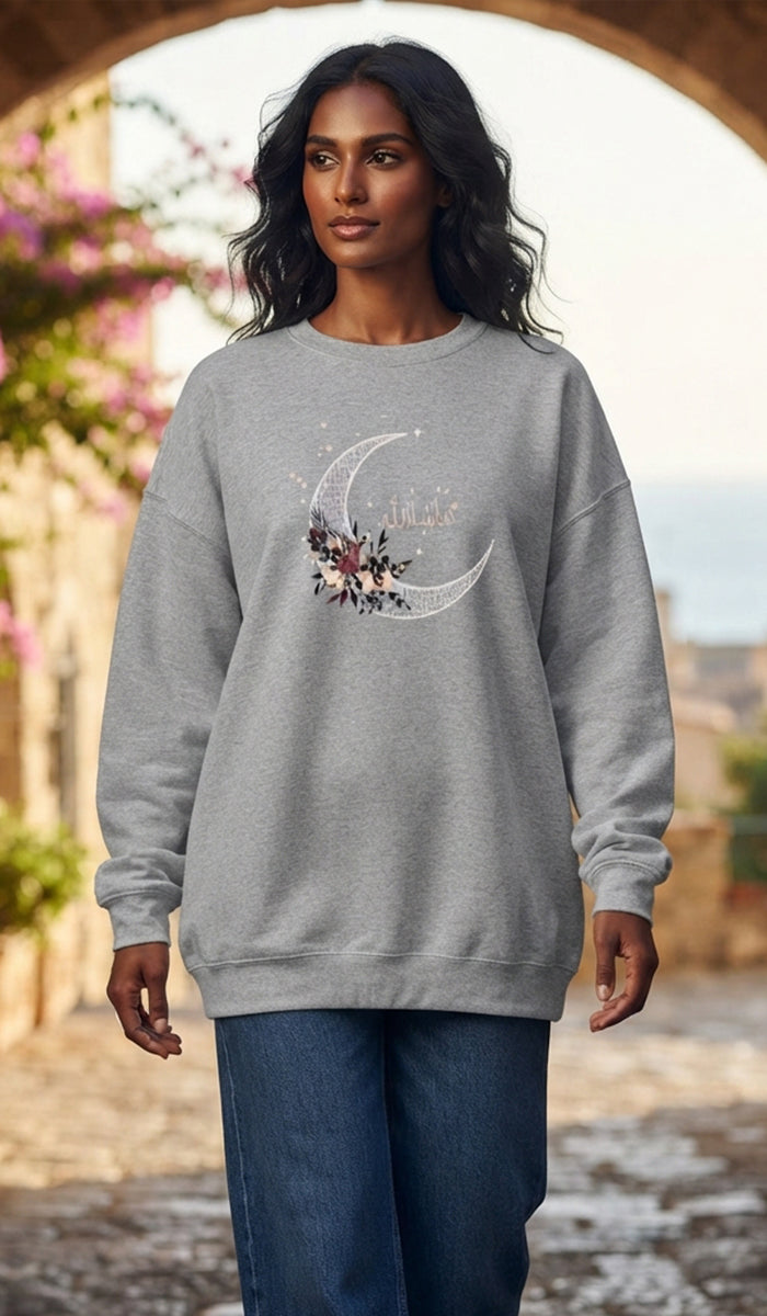 A woman wearing a light gray crewneck sweatshirt with a white crescent moon and floral graphic on the front, paired with blue jeans, standing in front of an outdoor stone archway.