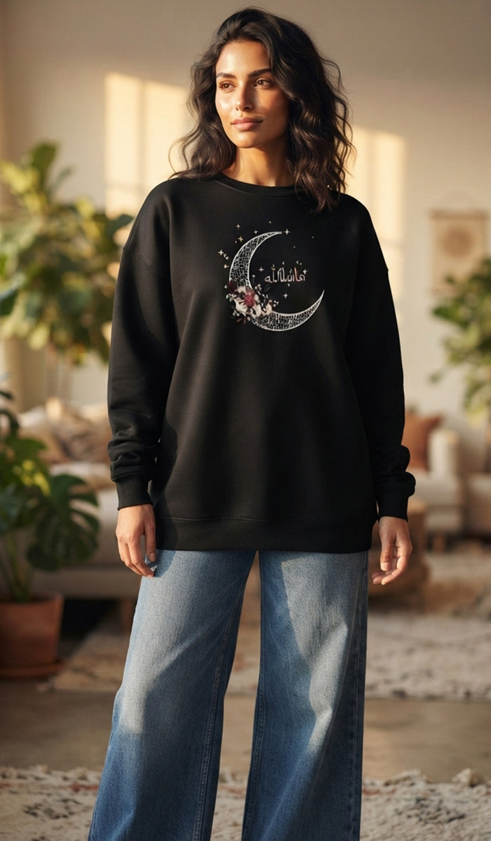 A woman wearing a black crewneck sweatshirt with a silver crescent moon and stars graphic on the front, paired with wide-leg blue jeans, standing in a softly lit indoor setting.
