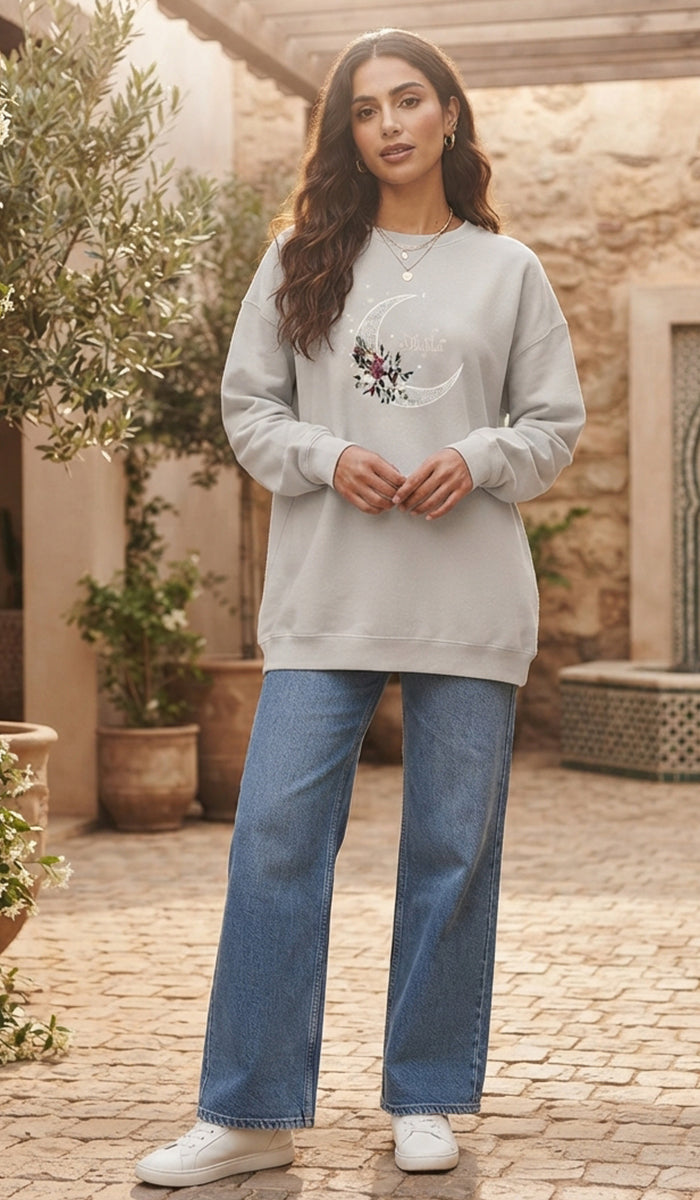 A woman stands outdoors wearing a light gray crewneck sweatshirt with a white crescent moon and floral graphic on the chest, paired with blue jeans and white sneakers.