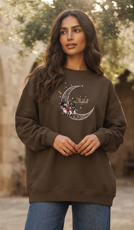 A woman wearing a dark brown crewneck sweatshirt with a crescent moon and floral graphic on the chest, paired with wide-leg blue jeans, standing in an outdoor stone courtyard.