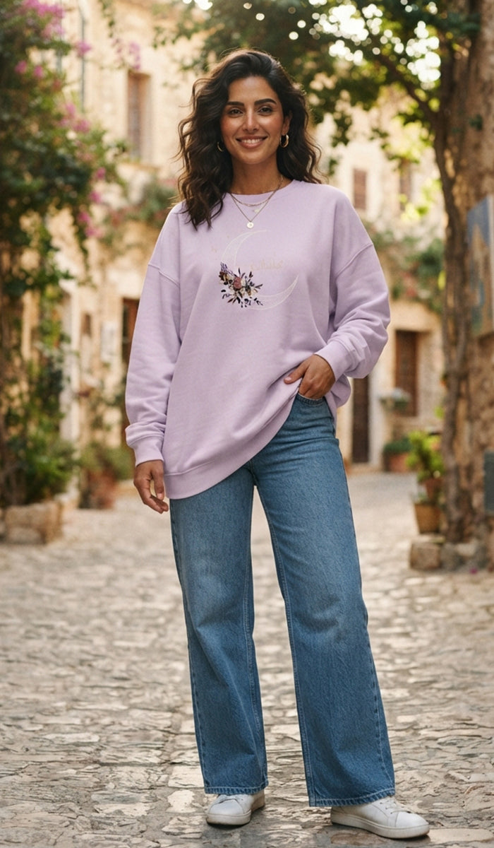 A woman stands on a cobblestone street wearing a lavender crewneck sweatshirt with a small floral graphic on the chest, paired with wide-leg blue jeans and white sneakers.