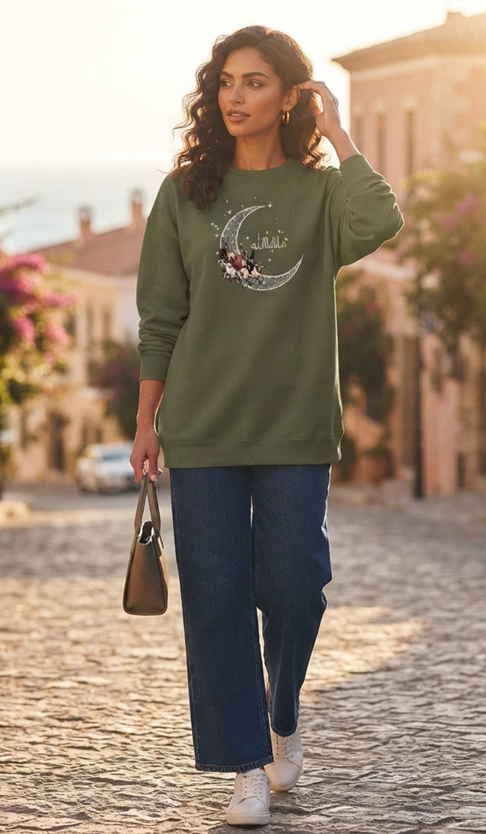A woman walking on a cobblestone street wearing an olive green long-sleeve sweatshirt with a moon and stars graphic and the word 'Galicia' across the chest, paired with wide-leg navy pants and white sneakers.