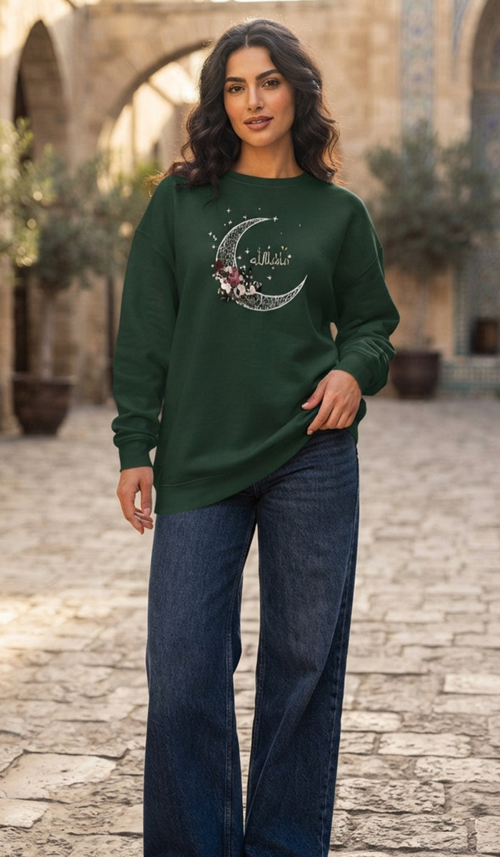 A woman wearing a dark green long-sleeve crewneck sweatshirt with a silver crescent moon and floral graphic on the chest, paired with dark blue jeans, standing in an outdoor stone courtyard.