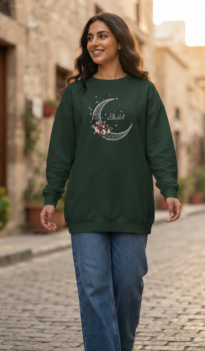 A woman wearing a dark green crewneck sweatshirt with a silver crescent moon and floral graphic on the chest, paired with blue jeans, walking down a cobblestone street.
