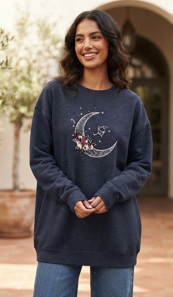 A woman wearing a navy blue crewneck sweatshirt with a silver crescent moon and floral graphic on the front, paired with blue jeans, standing in an outdoor courtyard setting.