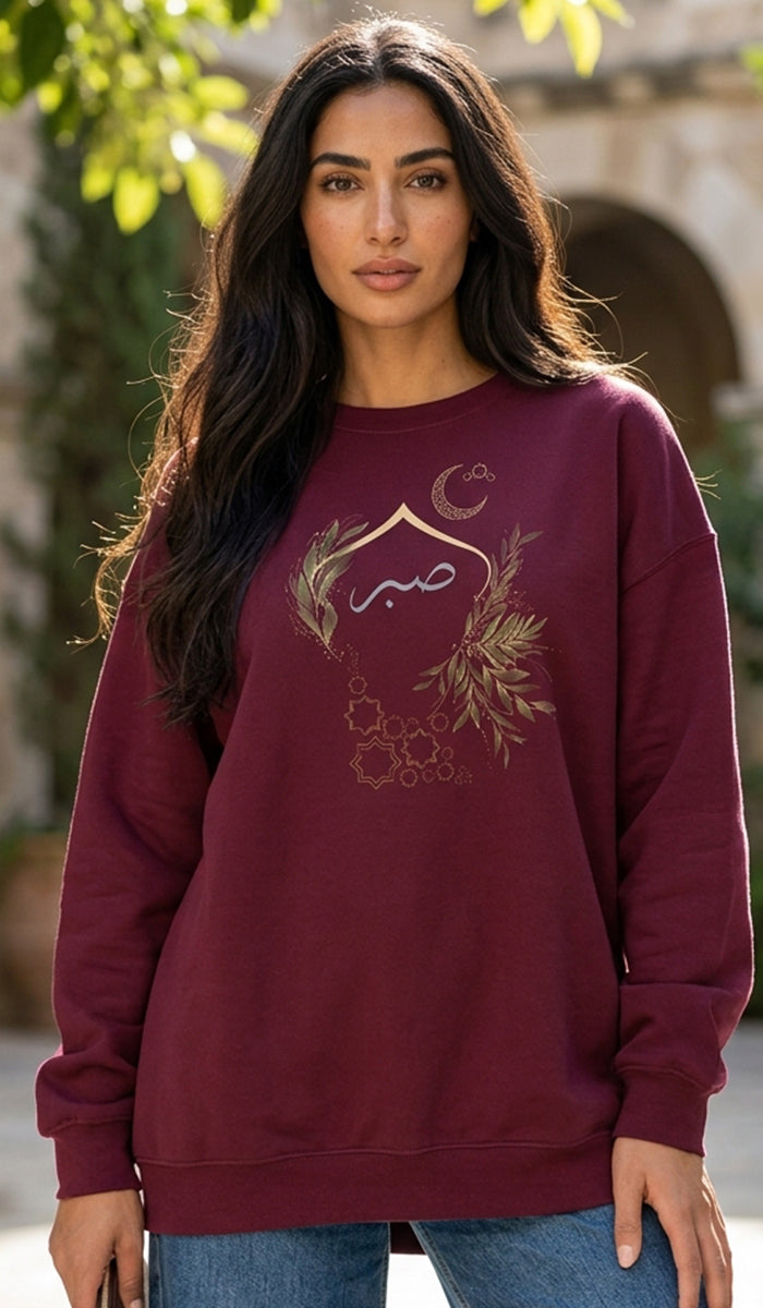 A woman wearing a maroon crewneck sweatshirt with a gold-colored Islamic graphic design on the front, paired with blue jeans, set against an outdoor background with soft-focus greenery.