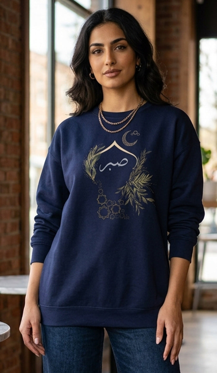 A woman wearing a navy blue crewneck sweatshirt with a gold Islamic graphic featuring a crescent moon, star, and Arabic calligraphy, paired with blue jeans in an indoor setting with brick walls.