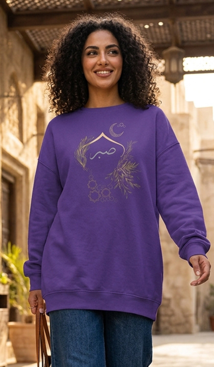 A woman with curly dark hair wearing a purple crewneck sweatshirt with a gold crescent moon and floral graphic on the front, paired with blue jeans, standing in an outdoor architectural setting.
