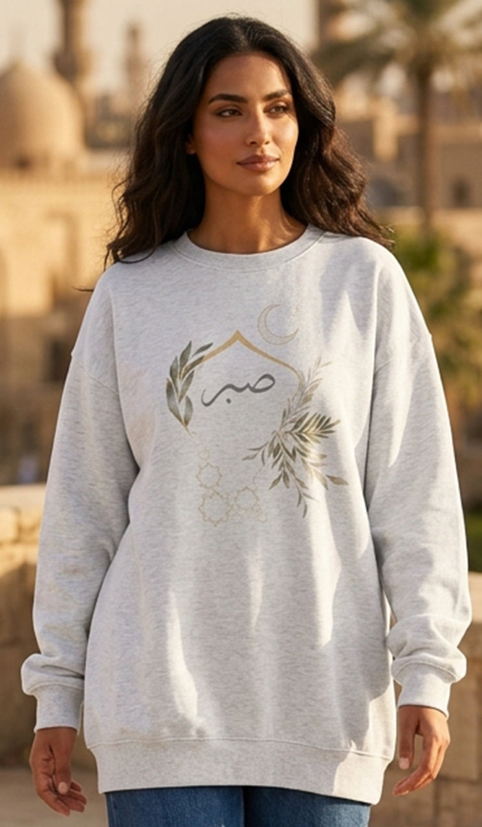 A woman wearing a light heather gray crewneck sweatshirt with a gold and green graphic of a crescent moon, stars, and floral elements, set against a blurred background of domes and palm trees.
