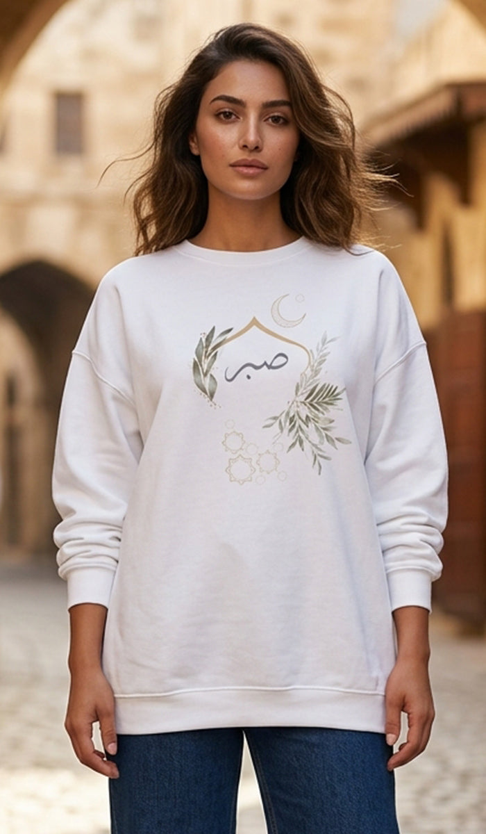 A woman wearing a white crewneck sweatshirt with a green and gold crescent moon and olive branch graphic on the front, paired with blue jeans, set against a blurred outdoor background of warm-toned stone buildings.