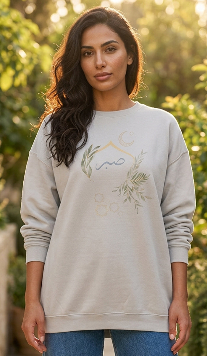 A woman wearing a light beige crewneck sweatshirt with a green and gold crescent moon and leaf graphic on the front, set against a blurred outdoor garden background.