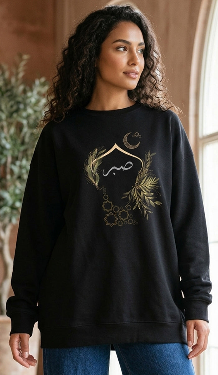 A woman wearing a black crewneck sweatshirt with a gold Islamic graphic featuring Arabic calligraphy, a crescent moon, and geometric patterns, paired with blue jeans.