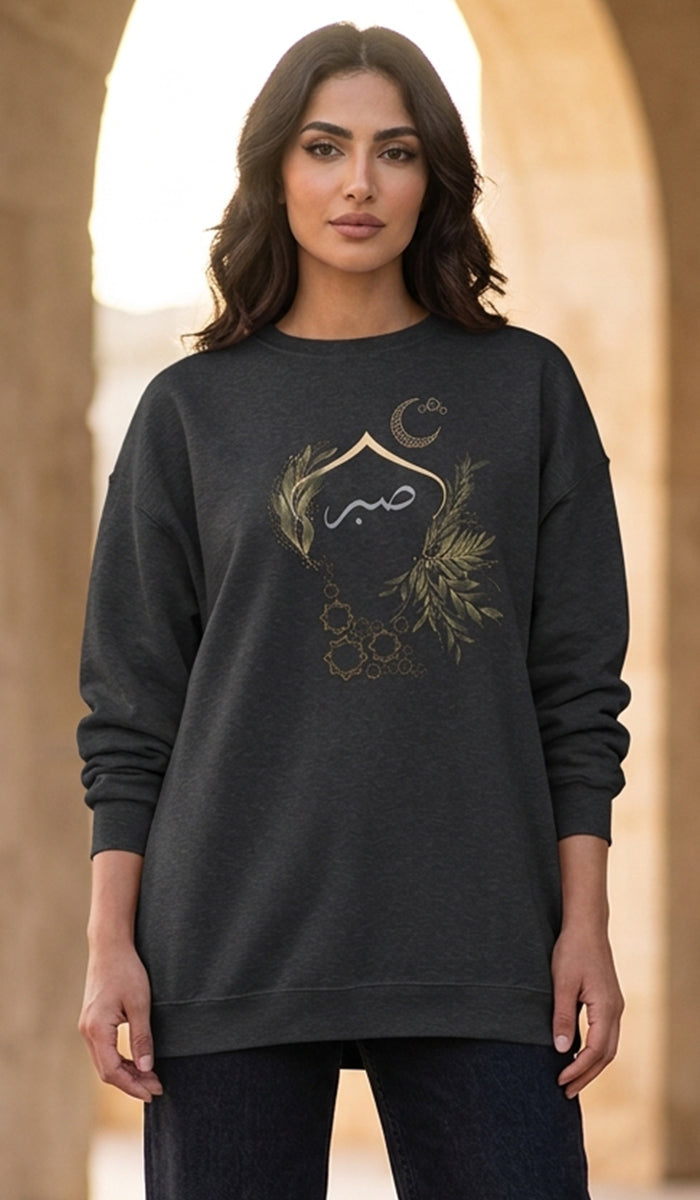 A woman wearing a dark charcoal gray crewneck sweatshirt with a gold and white graphic print featuring Arabic calligraphy, a crescent moon, and botanical elements, set against a blurred background of warm stone arches.