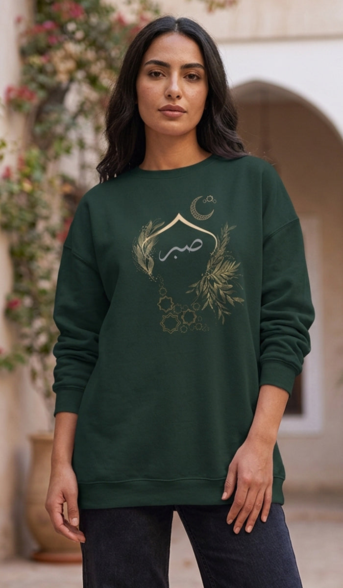 A woman wearing a dark green crewneck sweatshirt with a gold Islamic graphic featuring a mosque, crescent moon, and Arabic calligraphy, set against a blurred outdoor background.
