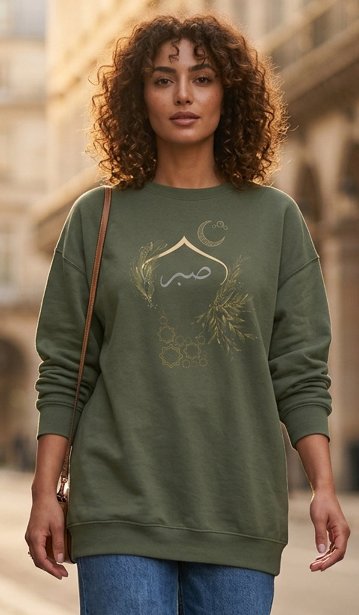 A woman with curly brown hair wearing an olive green crewneck sweatshirt with a gold crescent moon and floral graphic on the front, paired with blue jeans and a brown shoulder bag, set against a blurred outdoor architectural background.