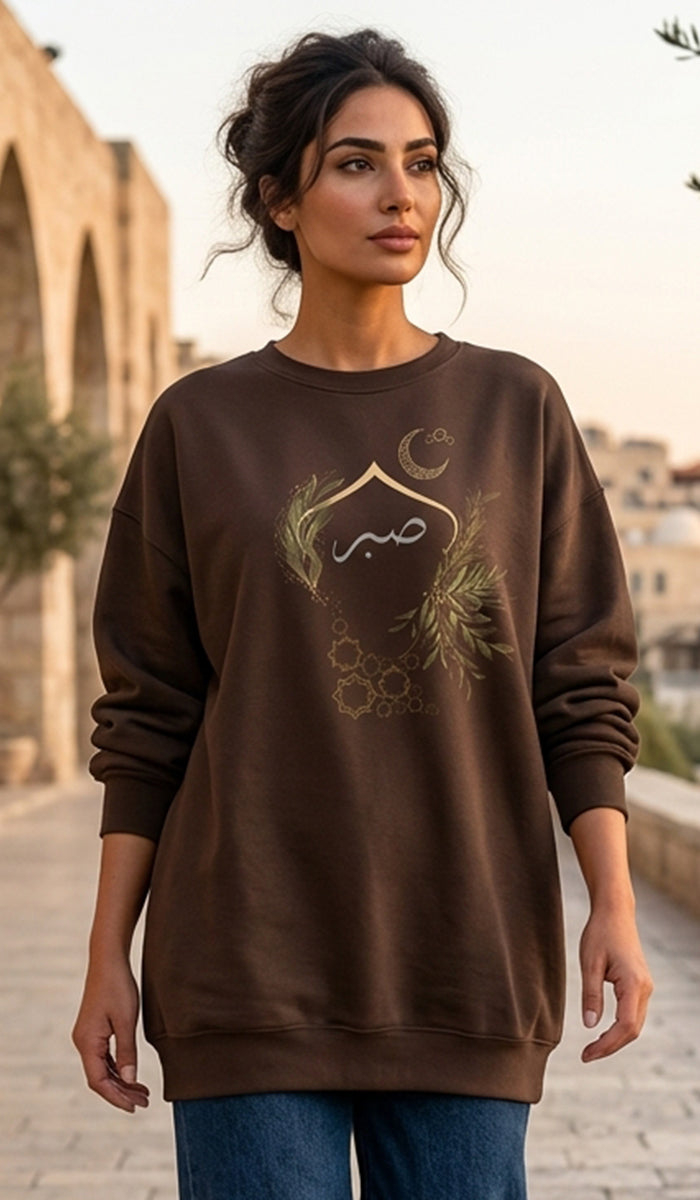 A woman wearing a brown crewneck sweatshirt with a gold crescent moon, mosque, and floral graphic on the front, standing outdoors in a sunlit urban setting.