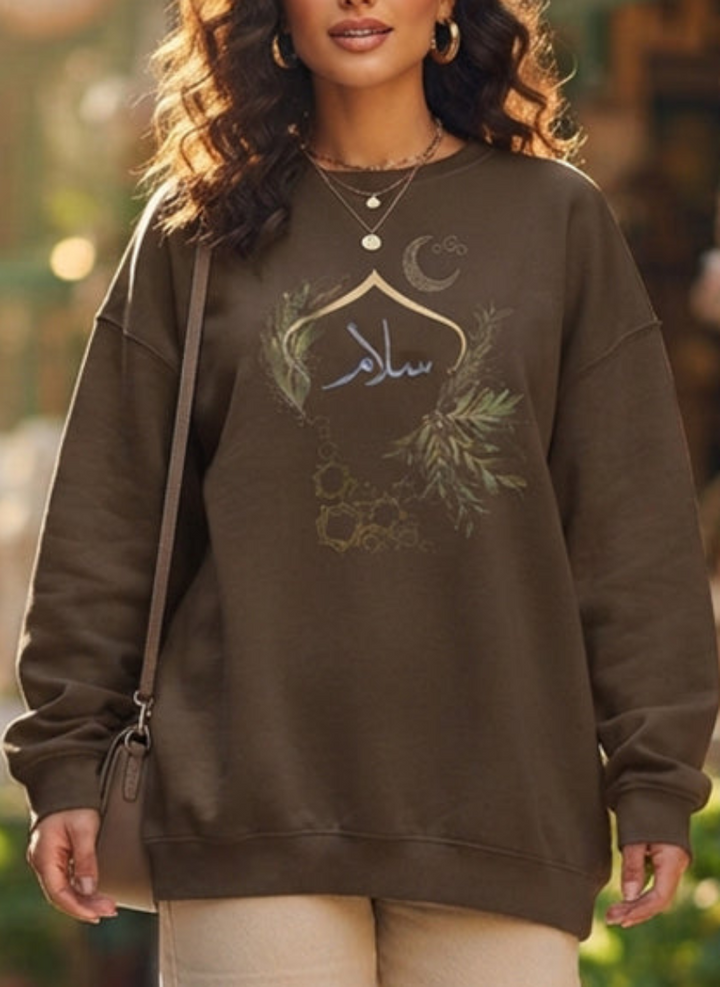 A woman wearing an oversized brown sweatshirt with a green and gold crescent moon and floral graphic on the front, paired with beige pants and a shoulder bag.