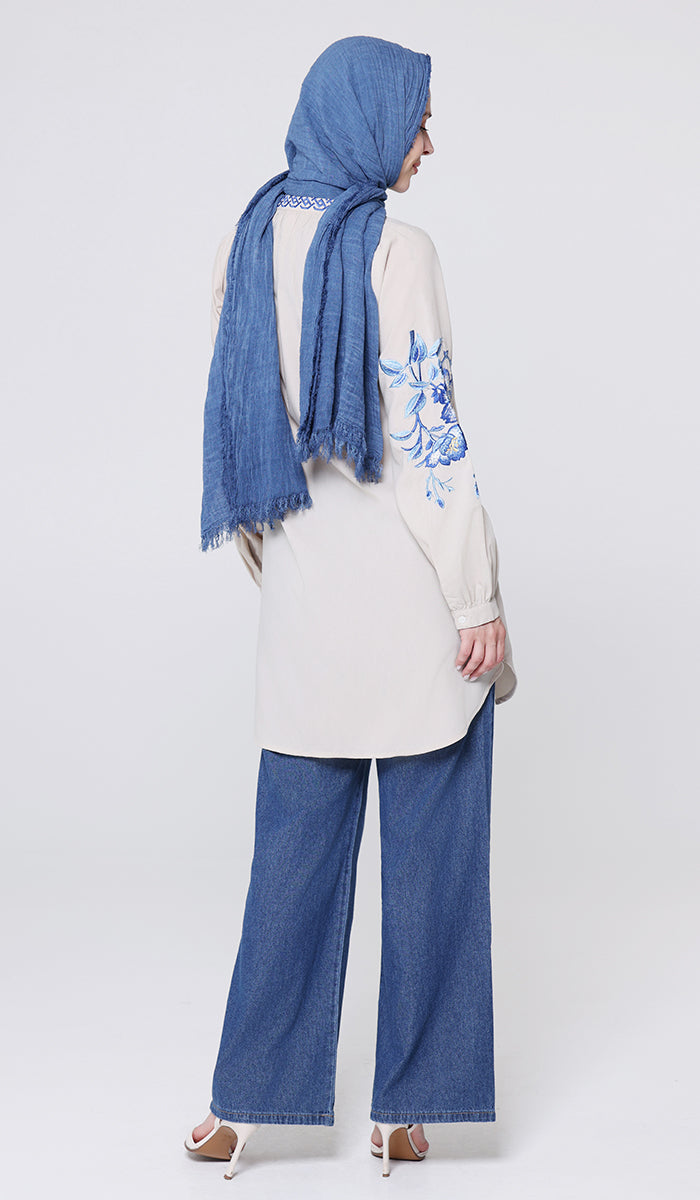 Woman wearing a blue hijab, white top with floral design, and blue jeans on a white background
