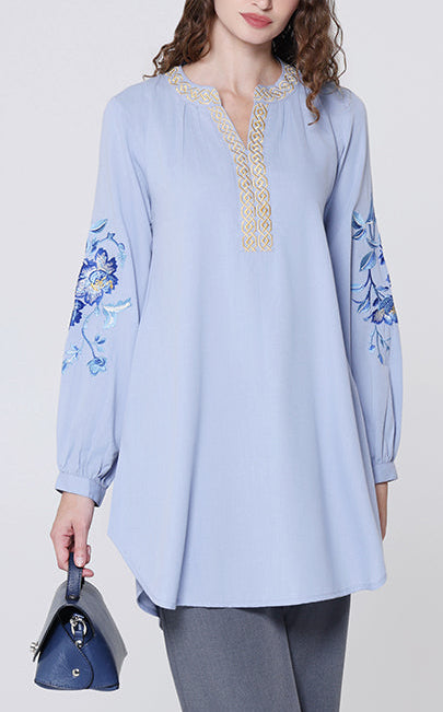 Woman wearing a light blue embroidered top and gray pants on a white background