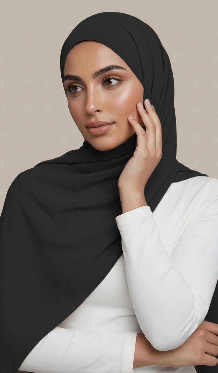 A woman wearing a solid black hijab draped elegantly over her shoulders and a white long-sleeve top, posed against a neutral beige background.