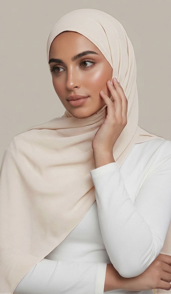 A woman wearing a light beige chiffon hijab and a white long-sleeve top, posing against a neutral beige background.