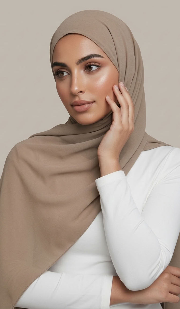 A woman wearing a taupe-beige hijab and a white long-sleeve top, posing against a solid light beige background.