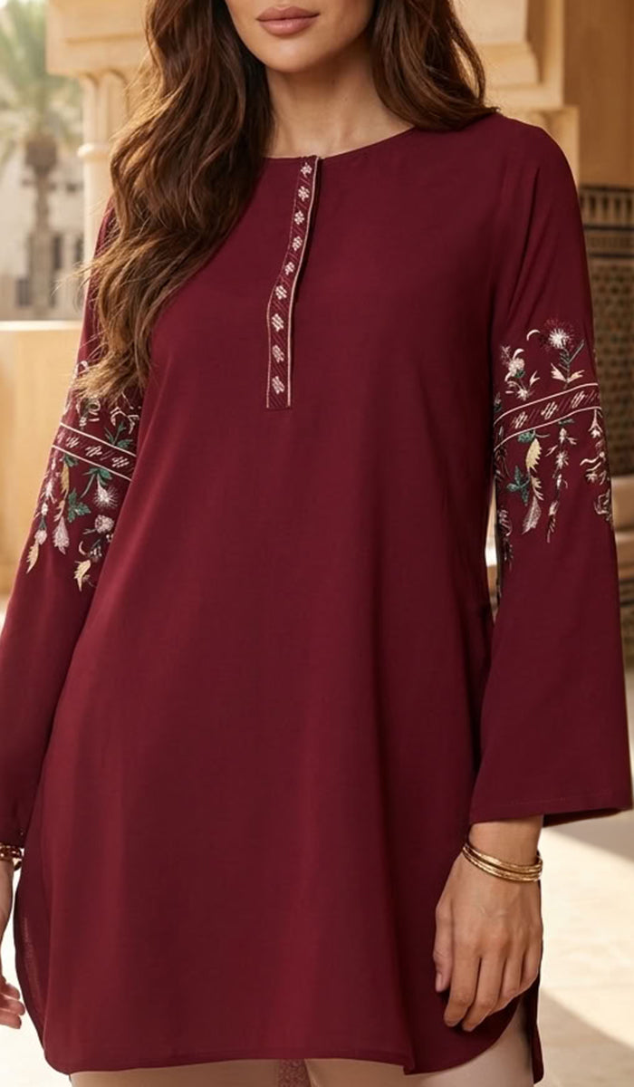 A woman wearing a long-sleeve maroon tunic dress with floral embroidery on the sleeves and a decorative embroidered neckline, shown in a warm-toned indoor setting.