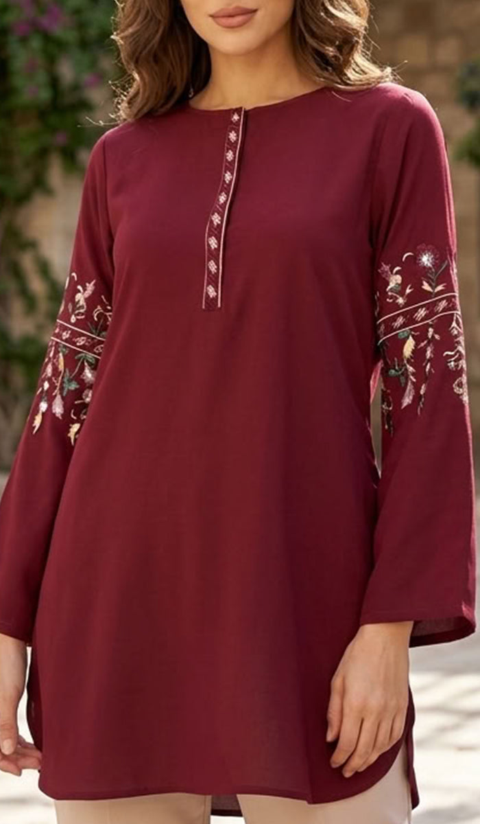 A woman wearing a long-sleeve maroon tunic top with floral embroidery on the sleeves and a decorative button placket at the neckline, shown in an outdoor setting.