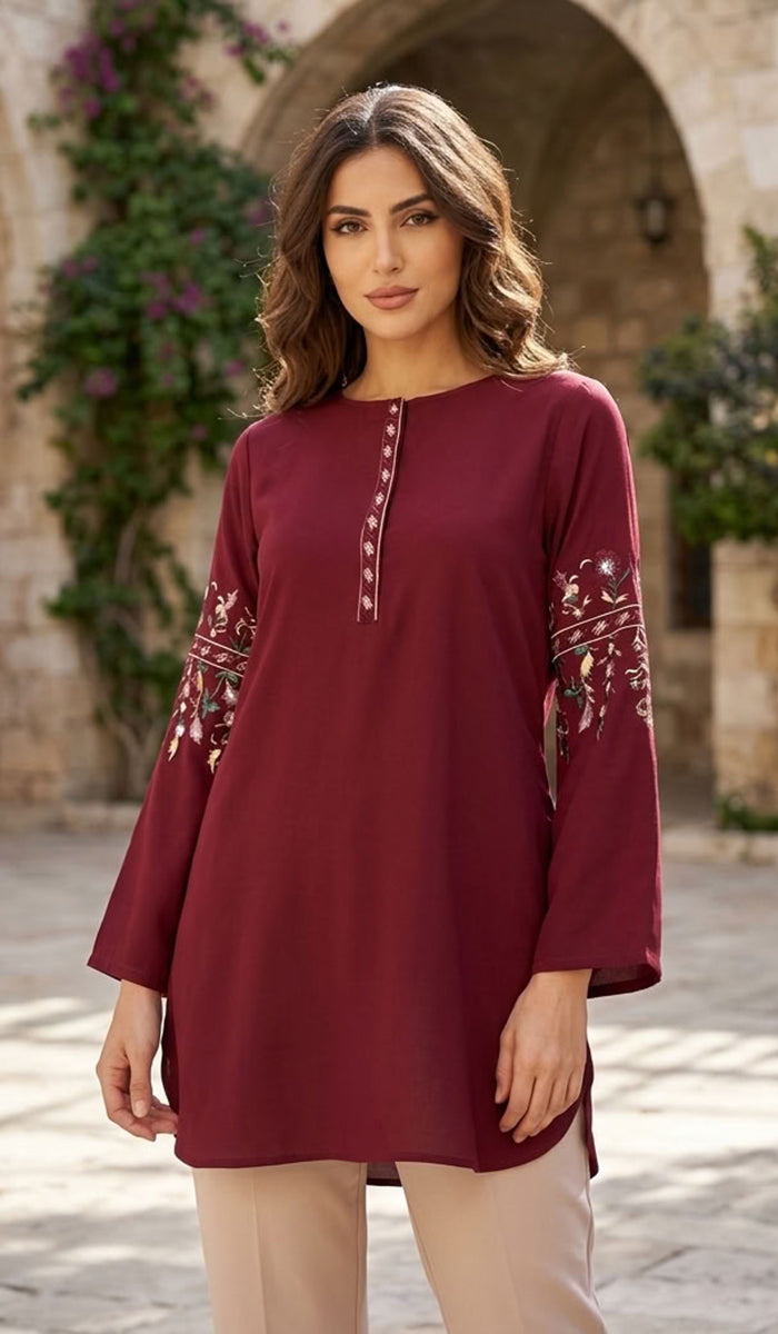 A woman wearing a long-sleeved maroon tunic top with white embroidery on the chest and sleeves, paired with beige trousers, standing in front of a stone archway.