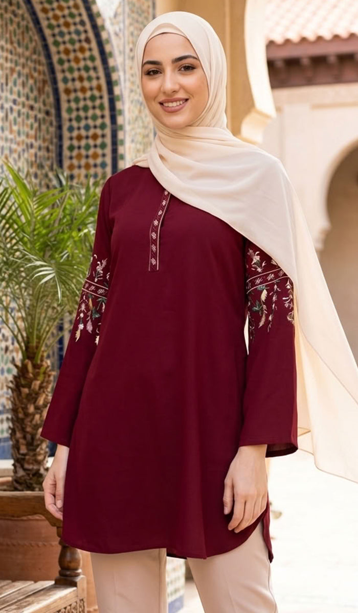 A woman wearing a deep maroon long kurta with floral embroidery on the sleeves and neckline, paired with beige trousers and a cream hijab, standing in front of a traditional Moroccan-style building with arches and a tiled wall.