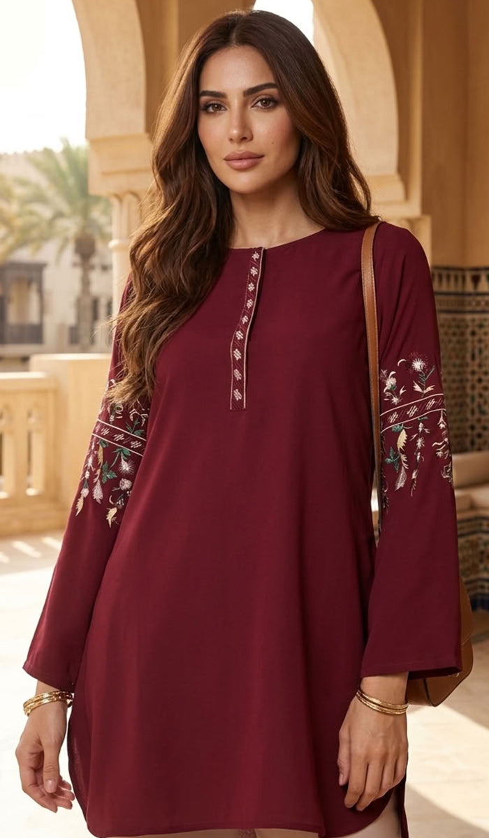 A woman wearing a long-sleeved maroon tunic dress with floral embroidery on the sleeves and a decorative neckline, standing in front of a warm-toned architectural background.