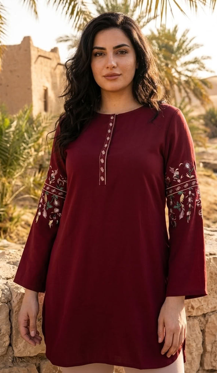 A woman wearing a long-sleeved maroon tunic with white and green floral embroidery on the sleeves and a matching embroidered neckline, standing outdoors in front of a stone wall and palm trees.