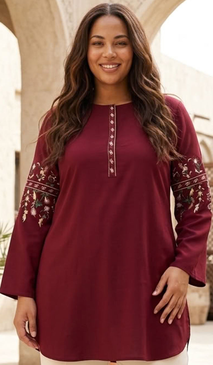 A woman wearing a long-sleeve maroon tunic top with embroidered details on the sleeves and a decorative button placket, set against a warm architectural background.